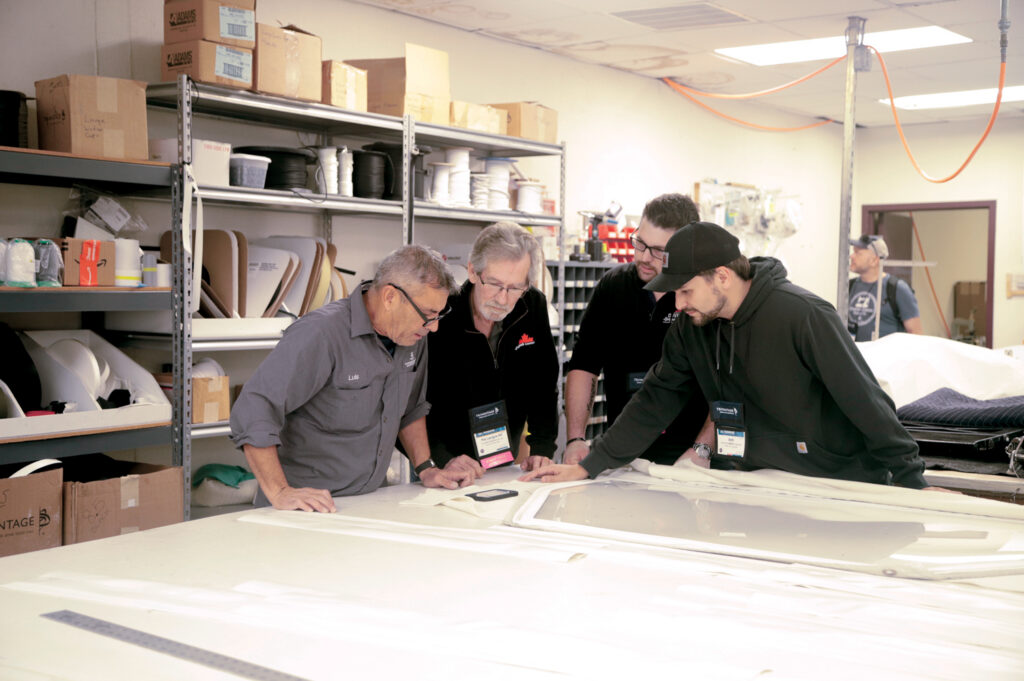 Four attendees examine materials on a table in a workshop setting, surrounded by shelves of supplies and tools, immersed in discussion.