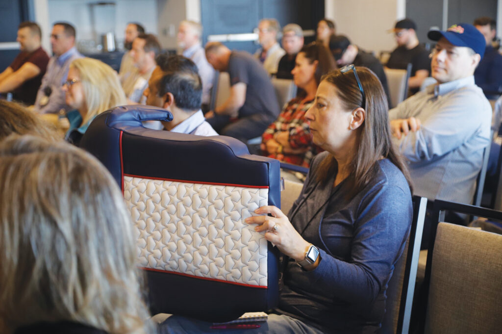A woman holds a patterned marine cushion while seated among an audience of attendees at a conference, featuring neutral-toned decor.
