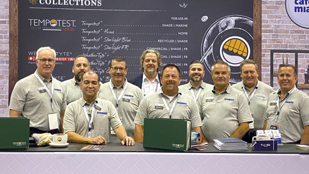 A group of ten men in gray shirts stand at a booth displaying fabric samples, with a large sign featuring "TEMPOST" and branding behind them.