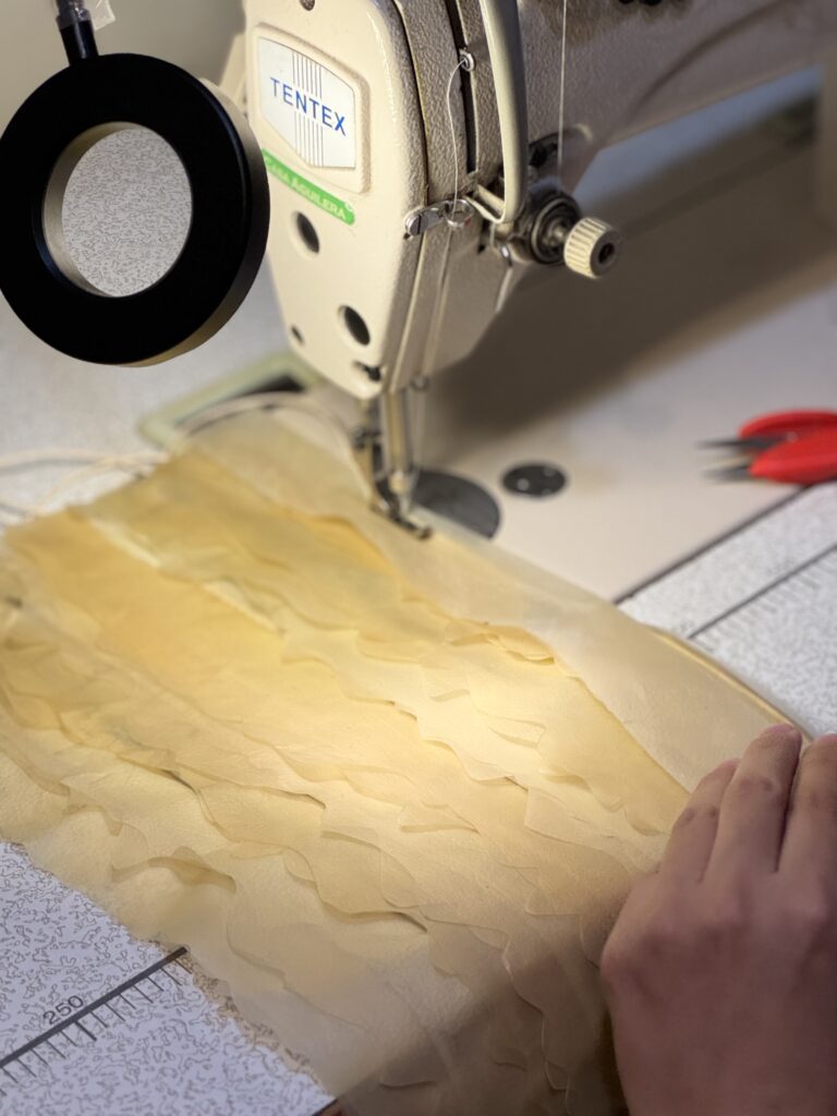A sewing machine with a magnifying glass hovers over layered translucent Celium bacterial cellulose fabric, while a hand guides the material on an industrial sewing machine table.