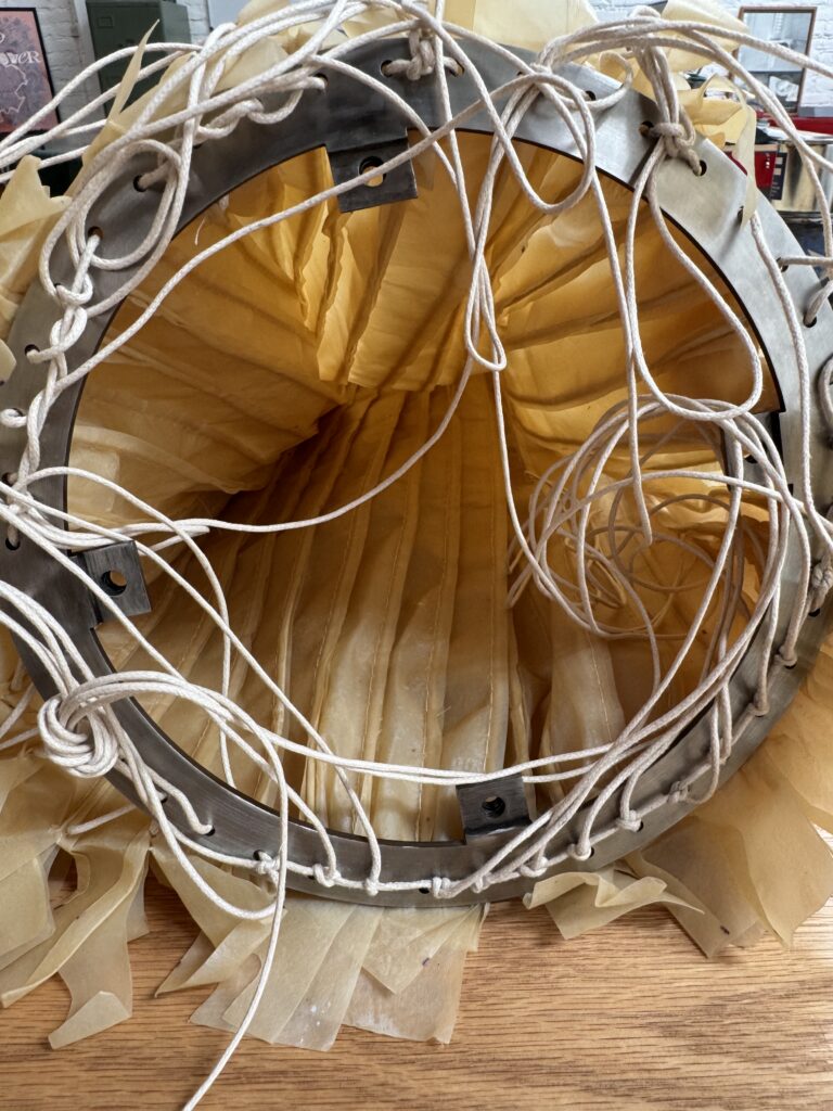 Interior view of a Polybion and Natural Urbano lamp structure with a circular metal frame, beige pleated bacterial cellulose Celium material, and intertwining ropes, resting on a wooden surface.
