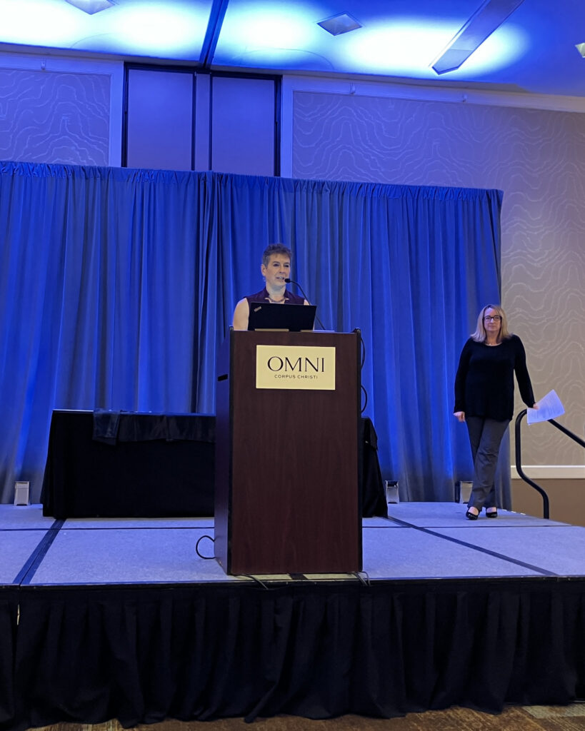 Charlene Clark, IFM, at a podium at the 2026 Marine Fabricators Association awards dinner. Behind her is a blue backdrop. She is engaged in presenting to an audience.
