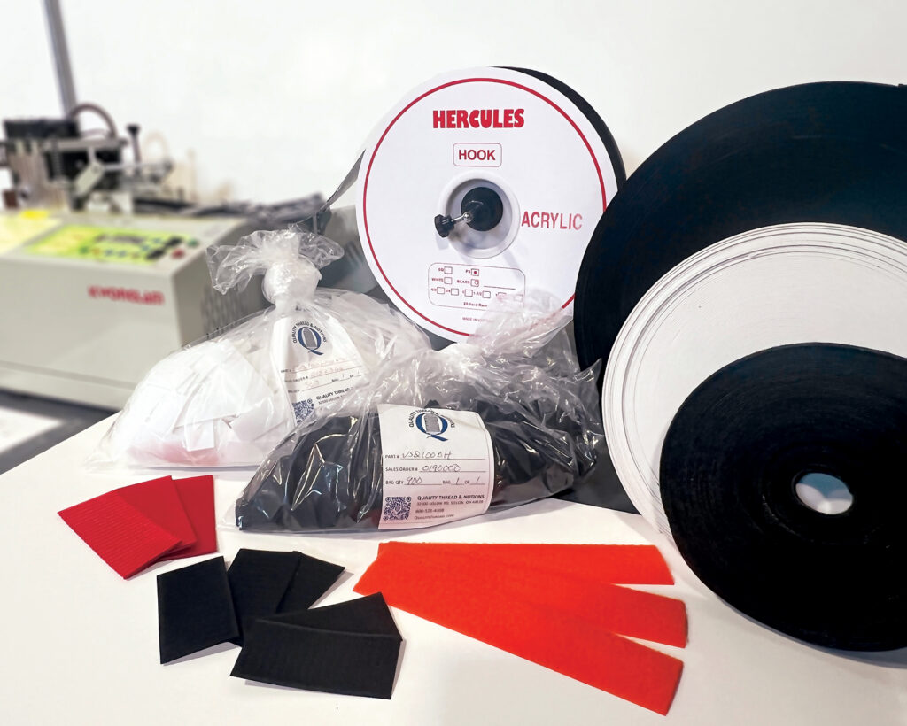 Assorted rolls, cut pieces and bagged orders of webbing, hook-and-loop and elastic in white, black, orange and red, arranged on a table with a machine in the background at Quality Thread & Notions. The company offers cutting services.