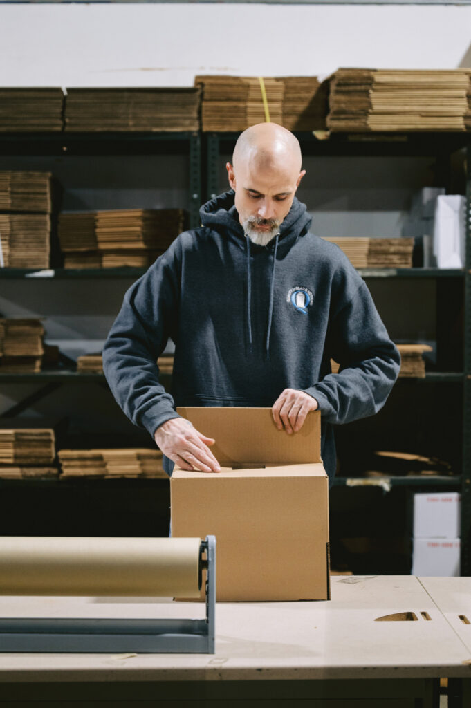 A male worker at Quality Thread & Notions in a dark hoodie folds a cardboard box on a table, with rolls of packing material in the foreground and shelves of boxes in the background.