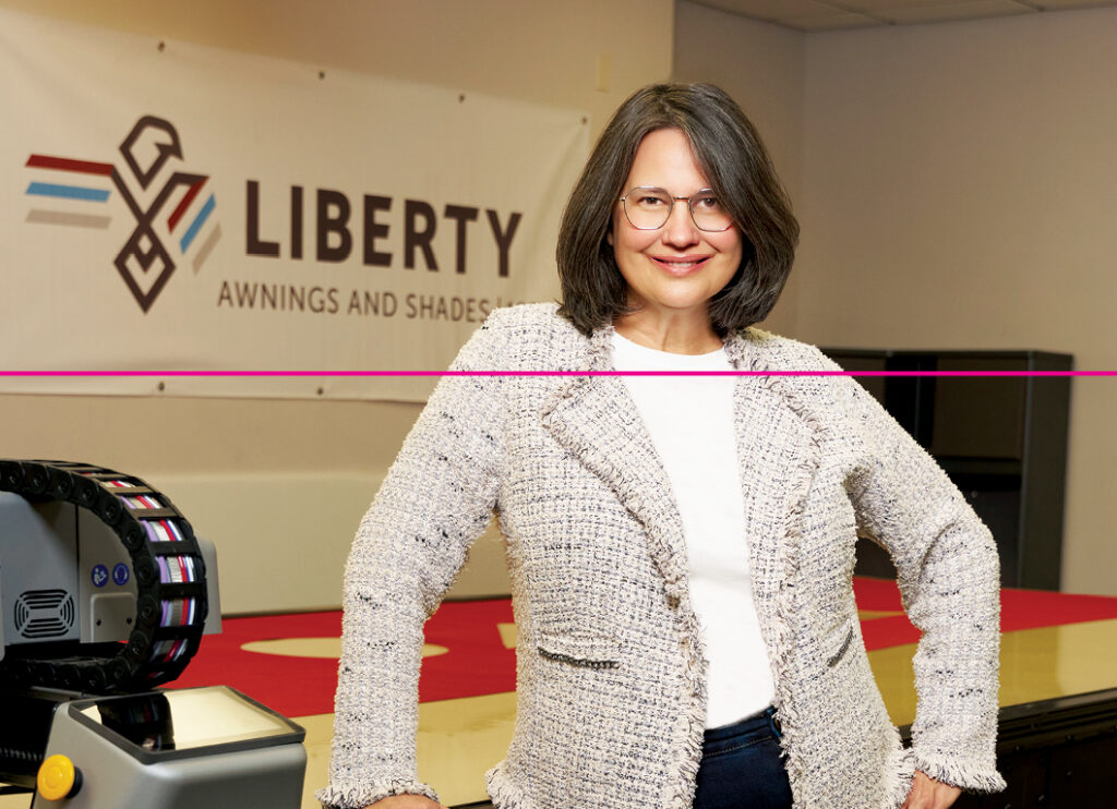 Woman with brown hair wearing a white jacket stands next to a table displaying red awnings, with a logo reading "LIBERTY" in the background.
