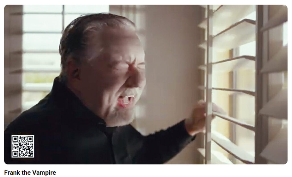 In Liberty Awnings and Shades commercial, Frank the Vampire, with graying hair and a dark shirt, grimaces dramatically at the sunlight coming through his slatted window blinds. A QR code is visible.