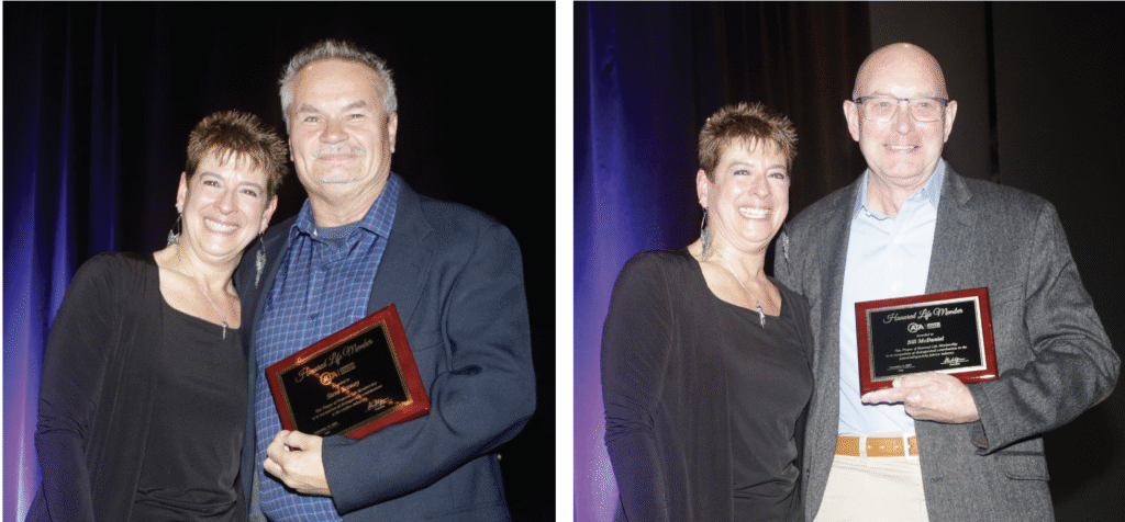 Two smiling individuals hold awards in a well-lit setting featuring dark curtains. The woman wears a black top, and the man a gray jacket over a blue shirt.