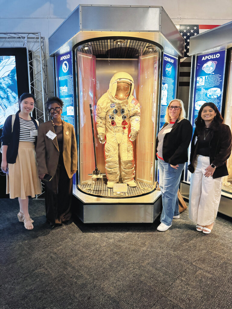 Four women pose together in front of a glass display featuring a NASA astronaut suit with protective seams, with informative panels in the background.