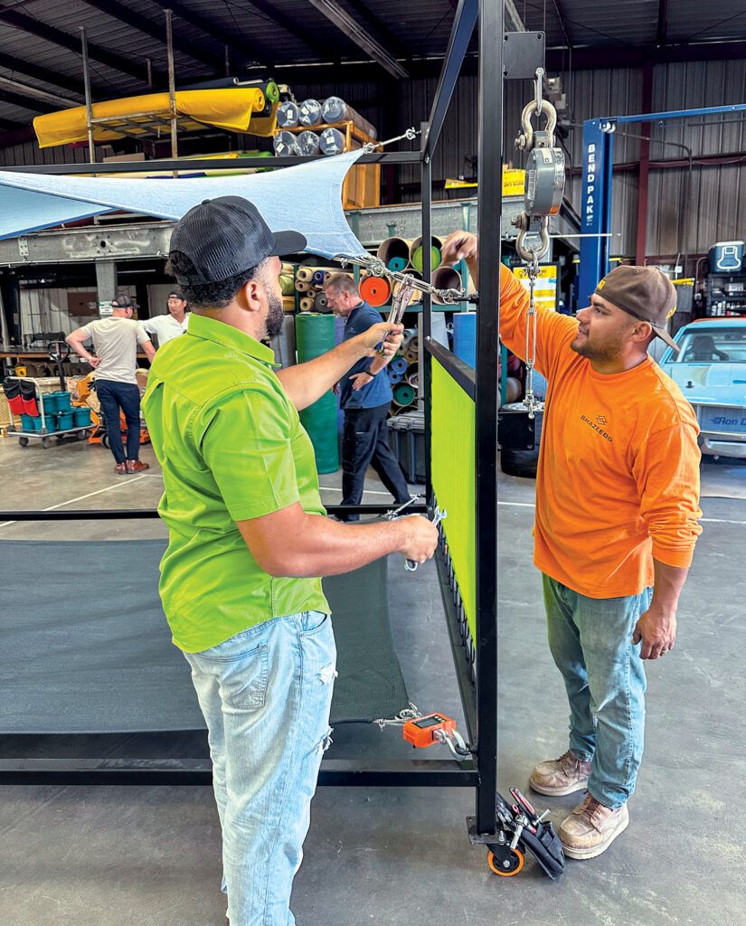 Two man in a warehouse adjust a blue fabric shade sail turnbuckle which is attached to a black metal frame. They are surrounded by colorful rolls of material and tools at Shade Sail University.