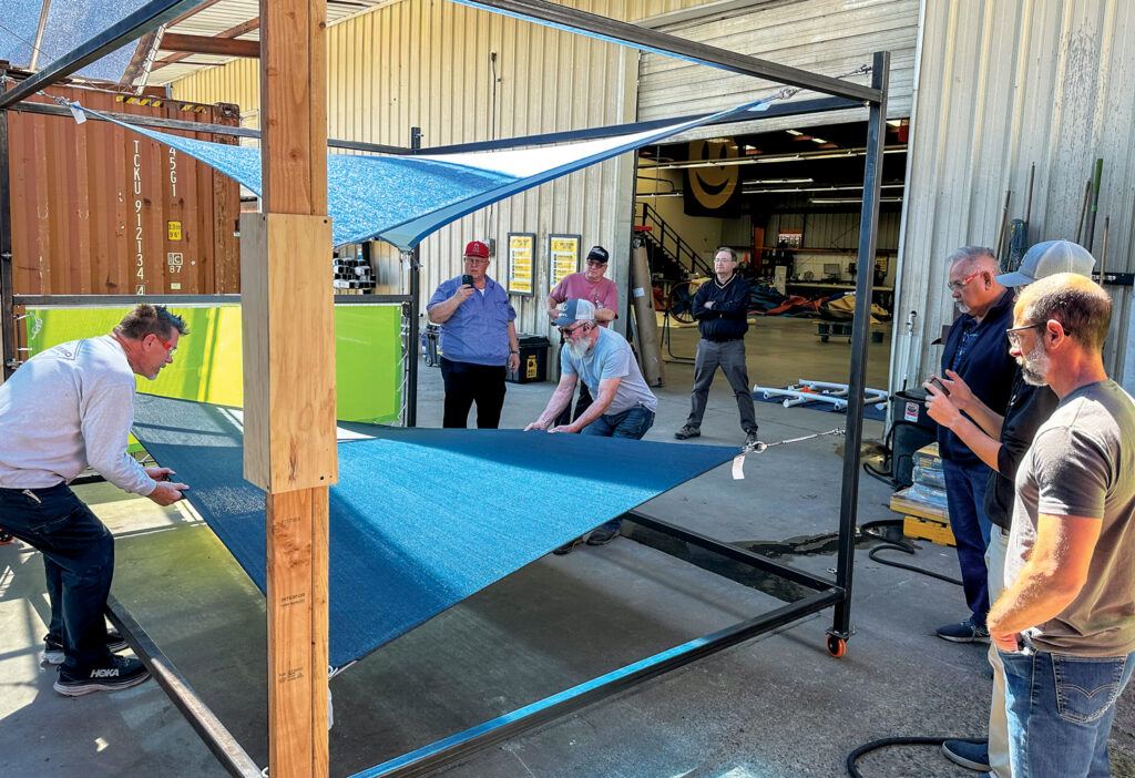 Five individuals observe as two men test a blue fabric shade sail stretched on a metal framework in a workshop setting at Shade Sail University.