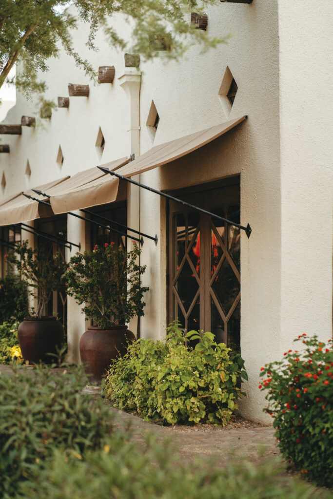 Tan awnings installed by Liberty Awnings and Shades at resort The Wigwam extend over tall windows that have decorative wooden accents on them. The windows are framed by large clay pots of greenery and blooming flowers against a textured white wall.