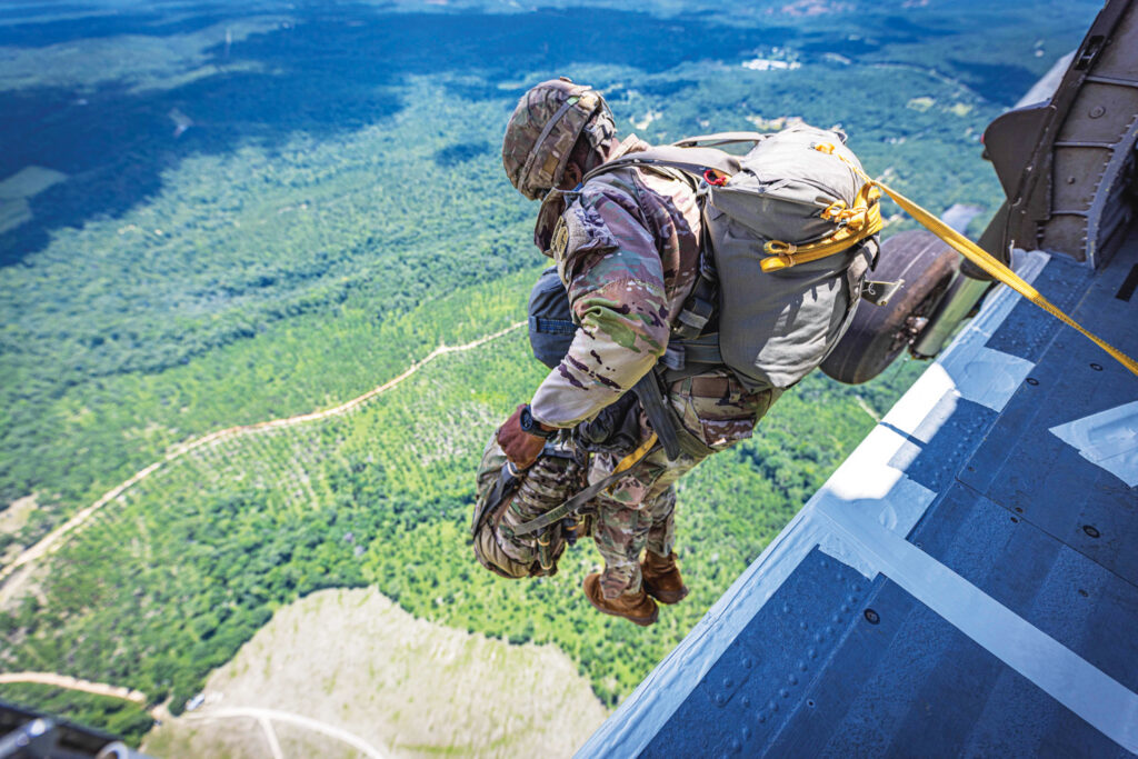 Soldier in camouflage gear prepares to descend from a helicopter, overlooking a lush green landscape and dirt paths below.