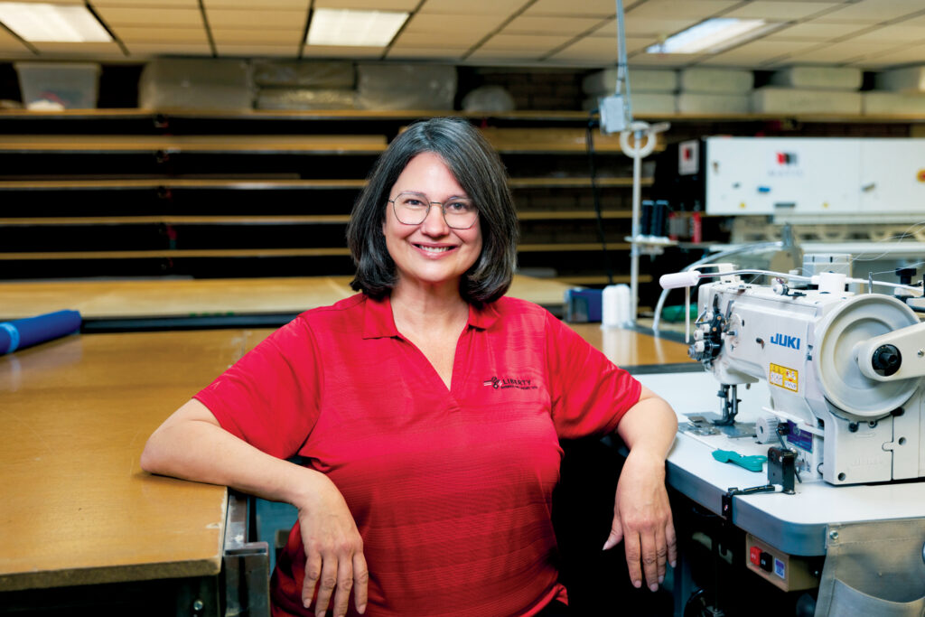 Amy Huette, with shoulder-length hair, smiles at the camera, wearing a red shirt in a workshop with sewing machines and fabric rolls in the background.