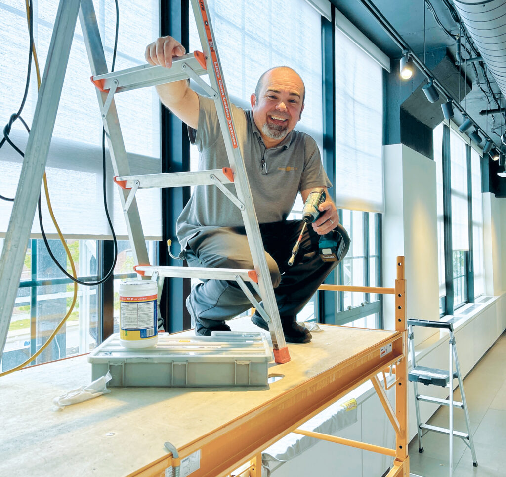 Company owner Roger Magalhães of Shades in Place on a scaffold installing window shades inside a commercial building, holding a drill with a ladder nearby, surrounded by large windows.