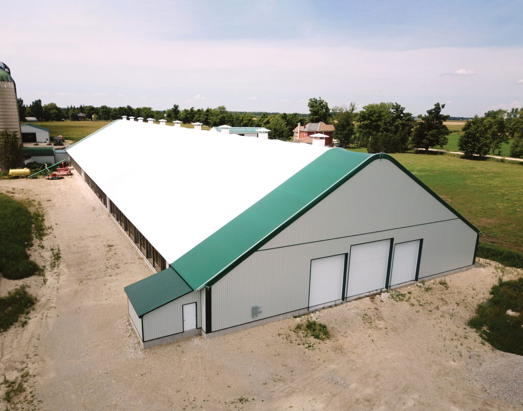 Large agricultural building with a white roof and green trim, surrounded by grass and trees under a clear blue sky.