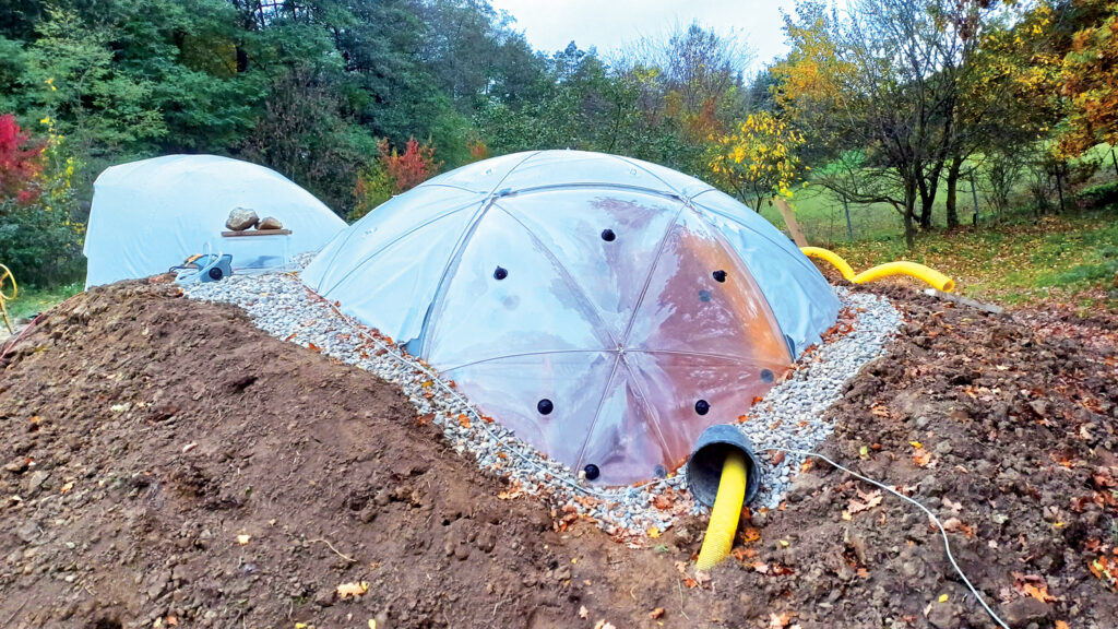 Pneumocell's geodesic dome structure, partially buried in soil, surrounded by gravel, with contrasting fall foliage in the background.