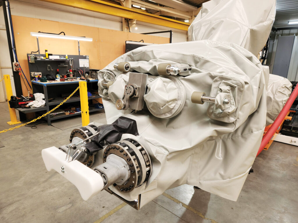 A robotic arm covered in beige protective skin, featuring mechanical joints and a white gripping attachment, set in a workshop environment at Kolorful Kanvas Ltd.