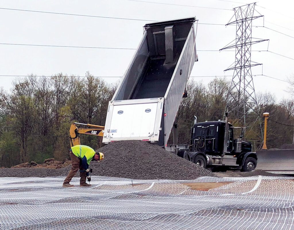 Construction site with a dump truck unloading gravel, while a worker in a yellow vest inspects Tensar's InterAx NX750 geogrid mesh on the ground.