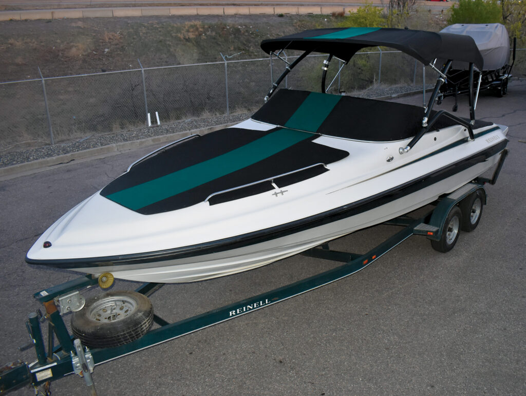 A sleek white boat with green and black striped covers and top, resting on a teal trailer against a gravel background. The project was done by Paul's Custom Canvas.