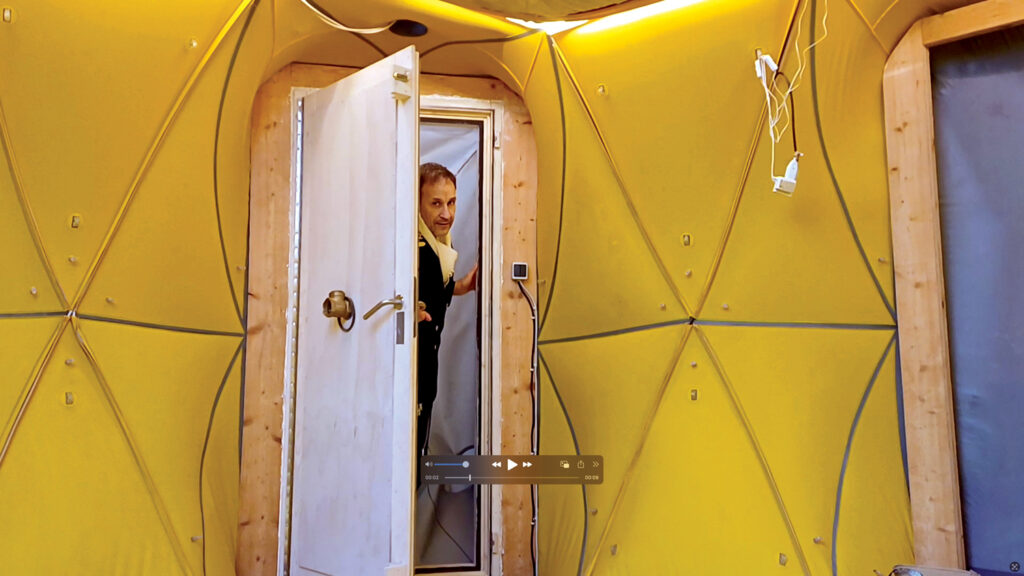 A person in a black jacket stands at an open door to an underground inflatable structure by Pneumocell, framed by a bright yellow, geometrically patterned interior.