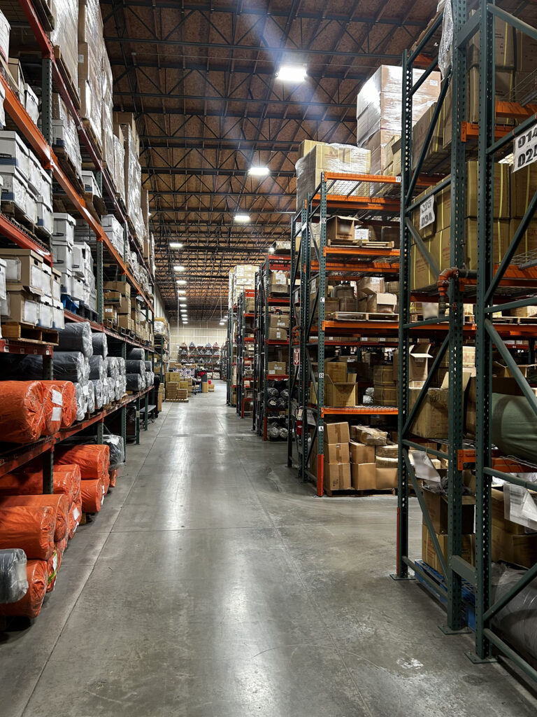 Spacious warehouse aisle lined with tall shelving units stocked with boxes and rolls of material, illuminated by overhead lights.