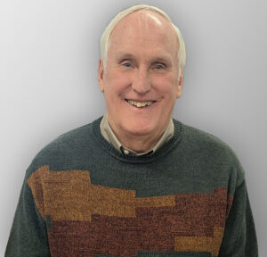 Dean Pecard of Laacke & Joys/LJ Design & Manufacturing, smiling. He is an older man with light hair, wearing a patterned green sweater over a collared shirt, against a soft grey background.