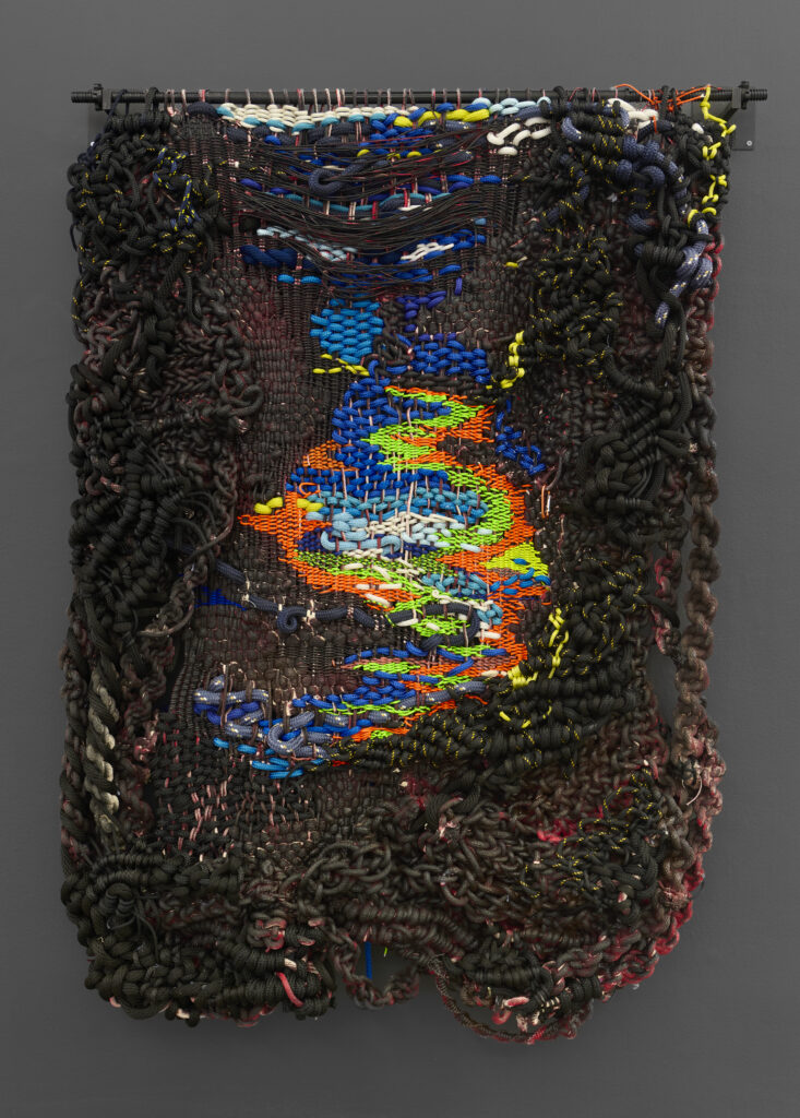 Colorful woven artwork hangs against a dark wall, featuring swirling patterns in bright blue, orange, green, and intricate textures.