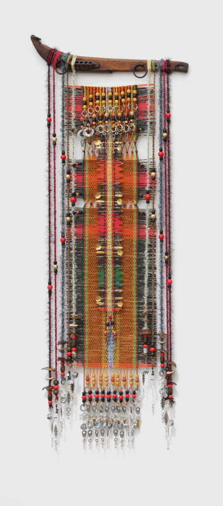 Colorful woven textile artwork features intricate patterns and embellishments, with hanging beads and metal accents. A wooden rod supports the piece.