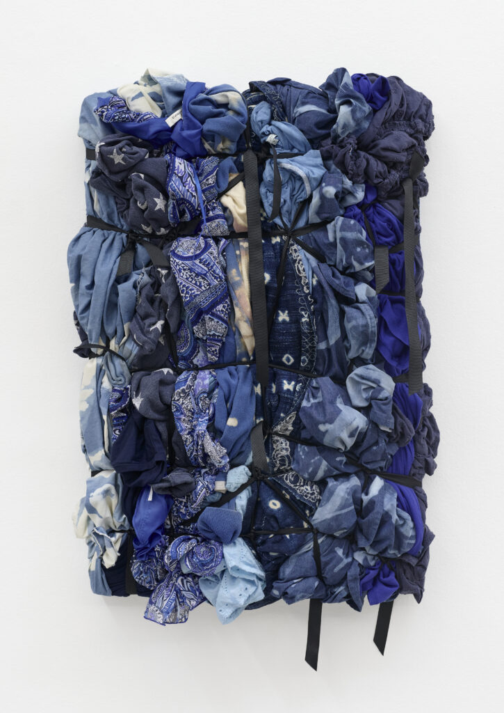 A textured arrangement of various blue fabrics, tied together with black straps, creating a colorful patchwork against a white backdrop.