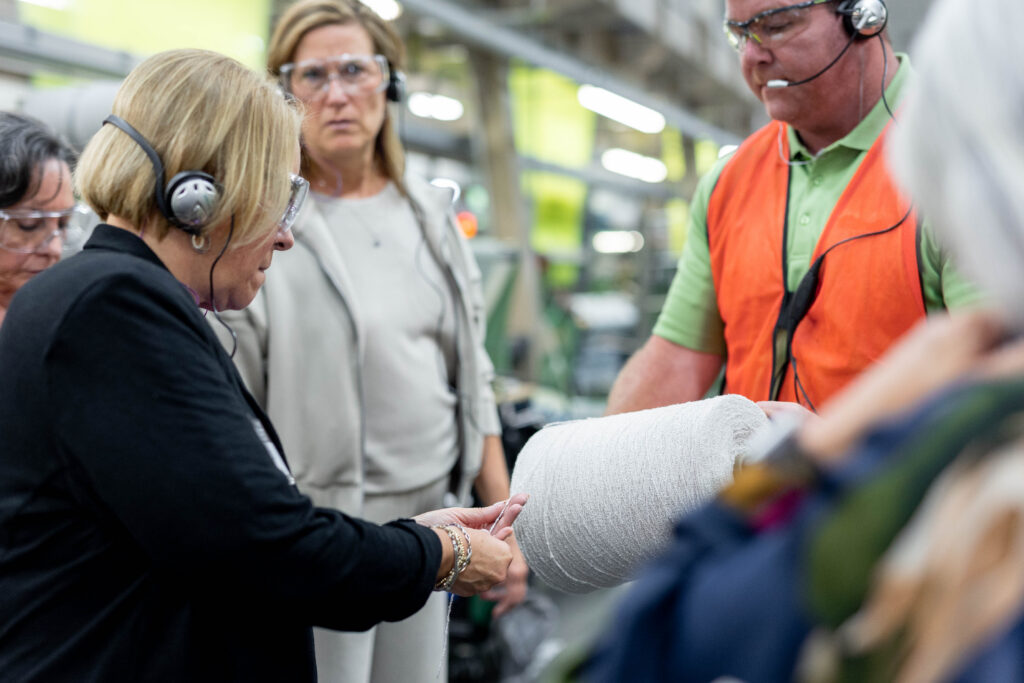 A group of people, wearing headsets and safety goggles, observes a man in an orange vest holding a roll of fabric in a factory setting.