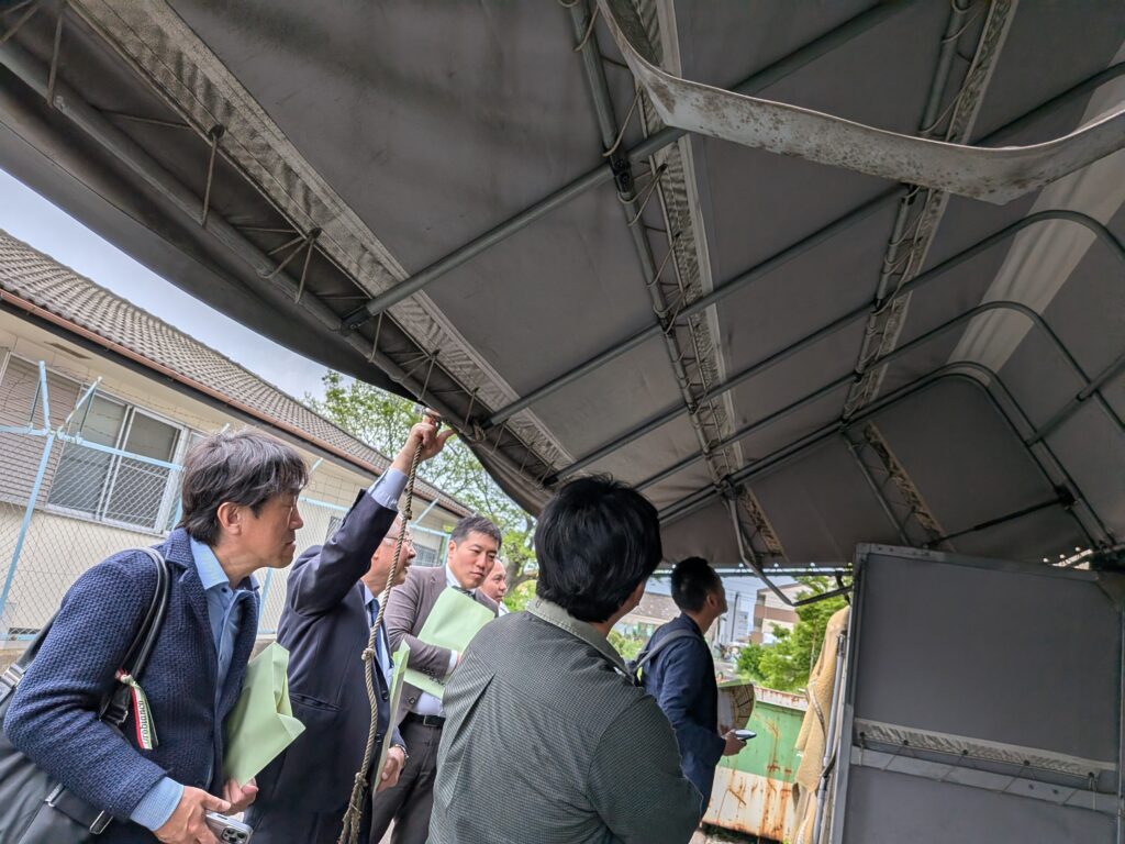 PXL_20260424_045315332 A group of men from ATA Japan, in formal attire, examines the interior of a large trailer of a wing-body truck system. The fabric truck cover is made by Avion Co. Ltd.