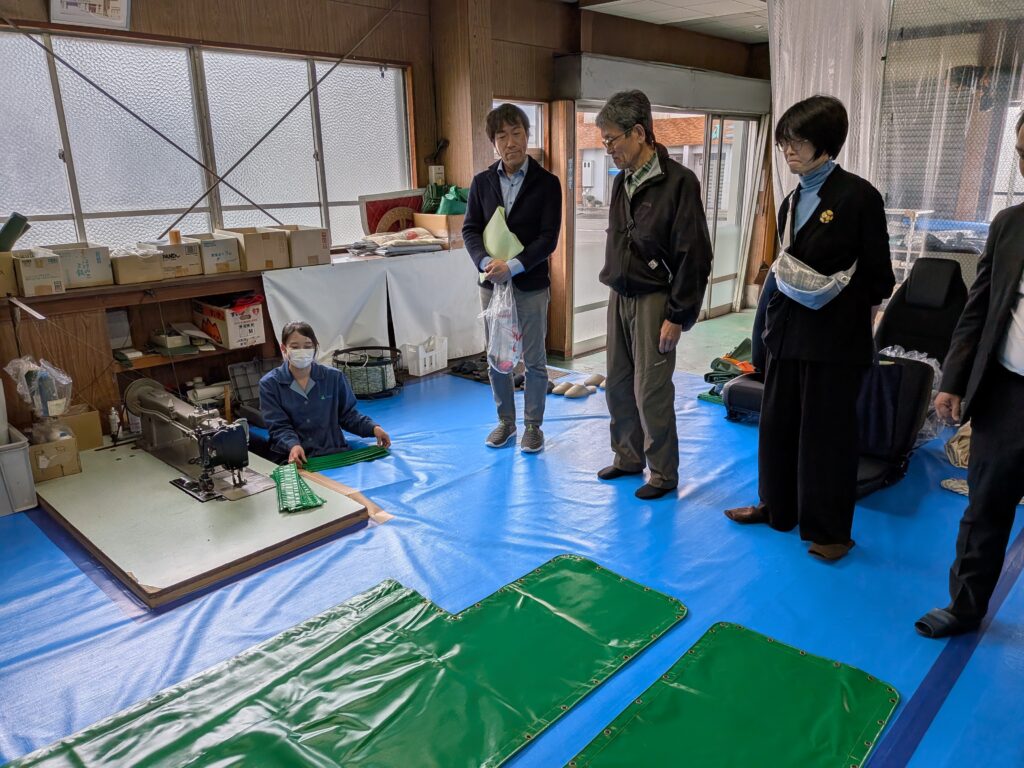 PXL_20260424_051724904.MP A group of four people from ATA Japan observes a woman seated recessed in a blue-covered floor, working on an industrial sewing project at Avion Co. Ltd. with long green fabric pieces in the foreground.