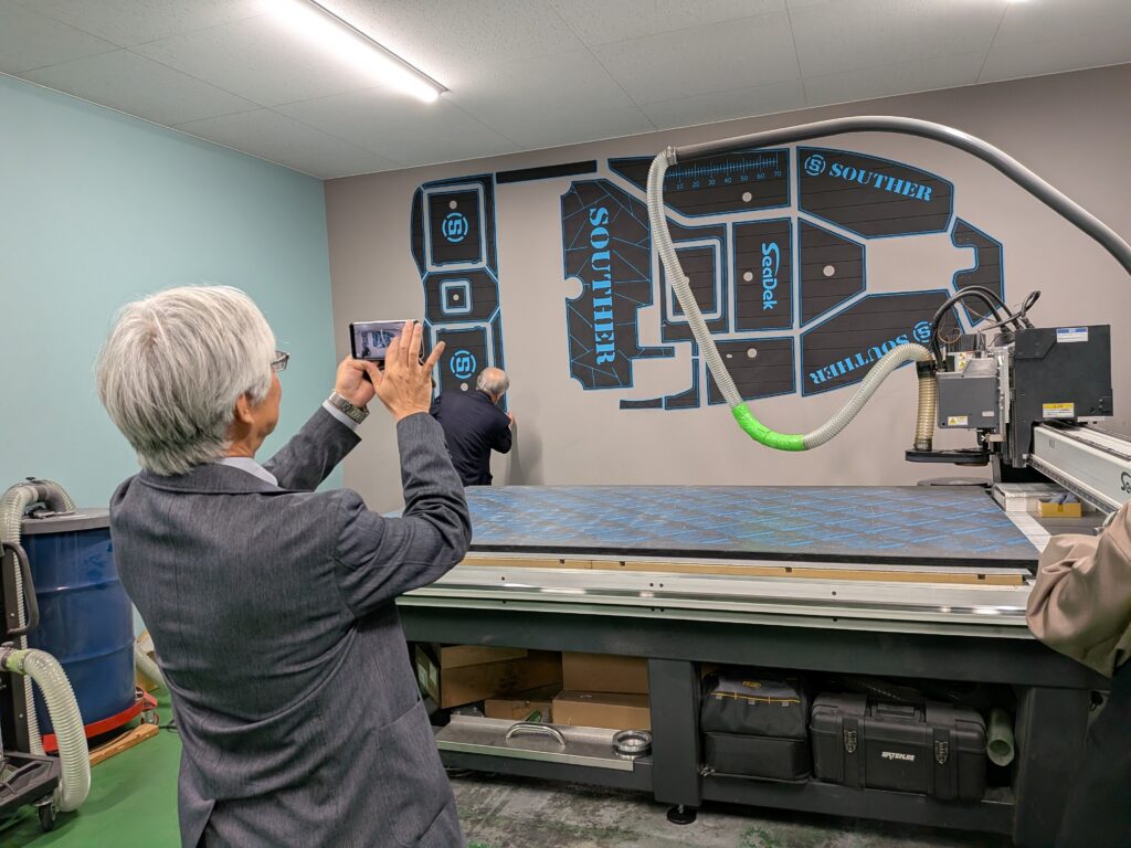 PXL_20260424_072830781.MP A man in a suit takes a photo of large blue and gray pattern pieces posted on the wall at Regar Marine Engineering Co. Ltd., behind an industrial cutting machine. The pattern pieces feature the words "SOUTHER" and "SeaDek."