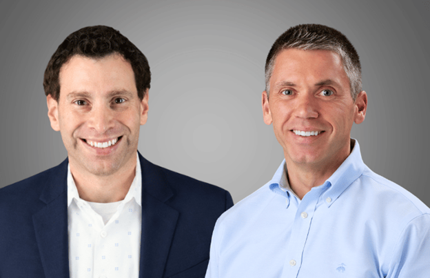 Two smiling men in professional attire, one in a dark blazer with a patterned shirt and the other in a light blue button-down shirt, against a gray background.