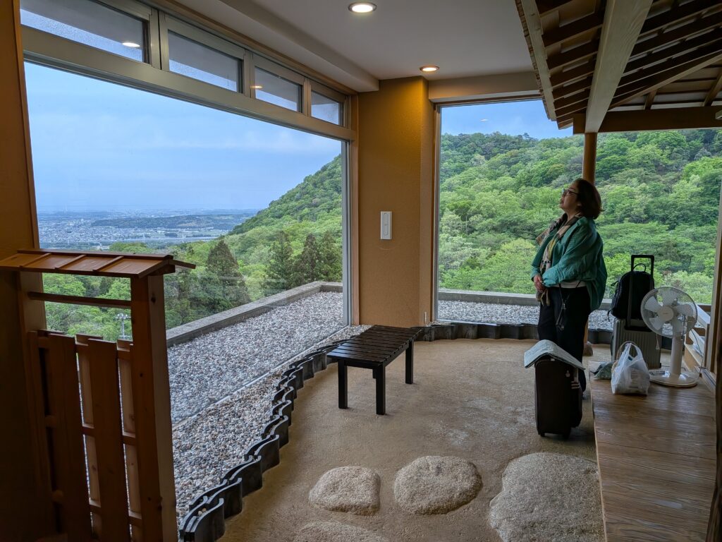 PresumablyHotSpringPXL_20260424_090750649.NIGHT A Japanese woman stands in a cozy room with large windows overlooking a lush green hillside at a hot spring resort in Japan. A suitcase and a small table are nearby.