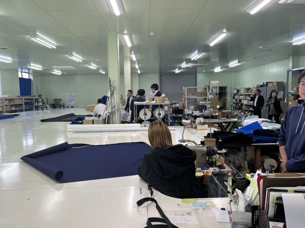 S__17891334_0 A spacious industrial textile fabrication workshop at Regar Marine Engineering Co. Ltd., with people working, featuring blue fabric rolls, sewing machines, and organized shelves filled with materials.