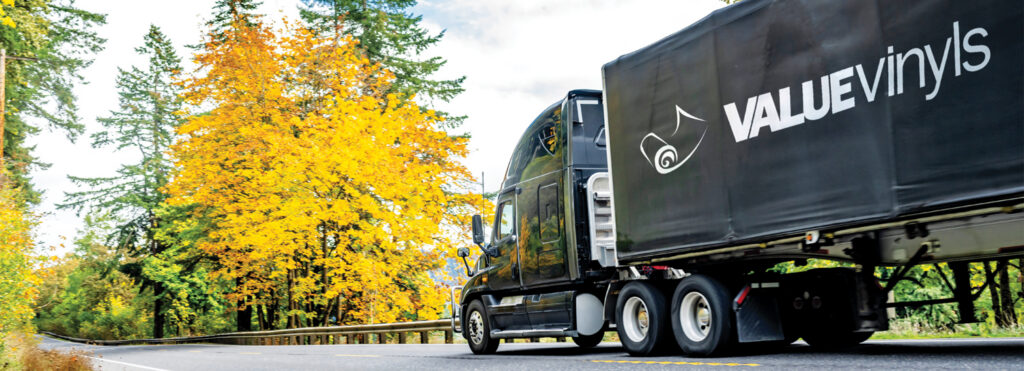 A black truck with a large "VALUE vinyls" logo on a Tempo 680 fabric truck cover system drives along a road lined with vibrant yellow and green autumn trees.