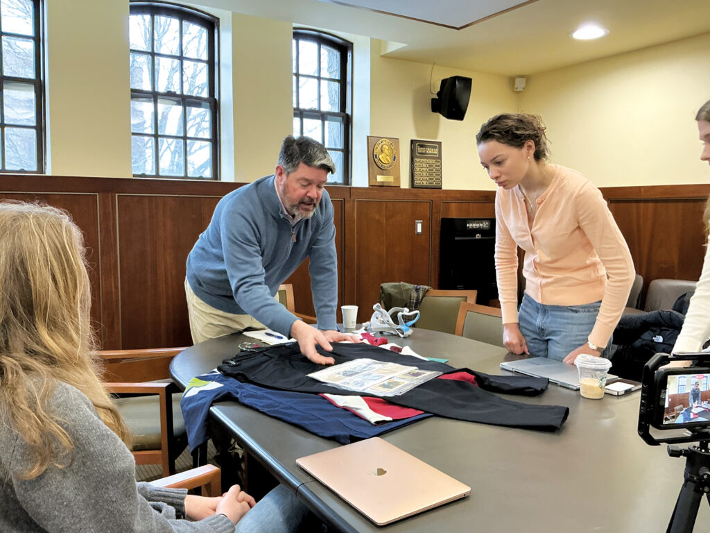 A man demonstrates medical textiles on a table, while three women observe. The setting features wooden paneling and large windows.
