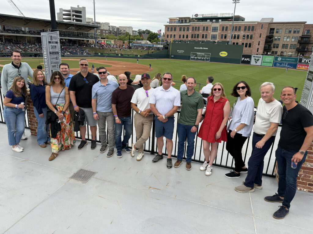 Group of professionals posing in front of a baseball field, with a mix of casual attire and vibrant backgrounds, enjoying an outdoor event.
