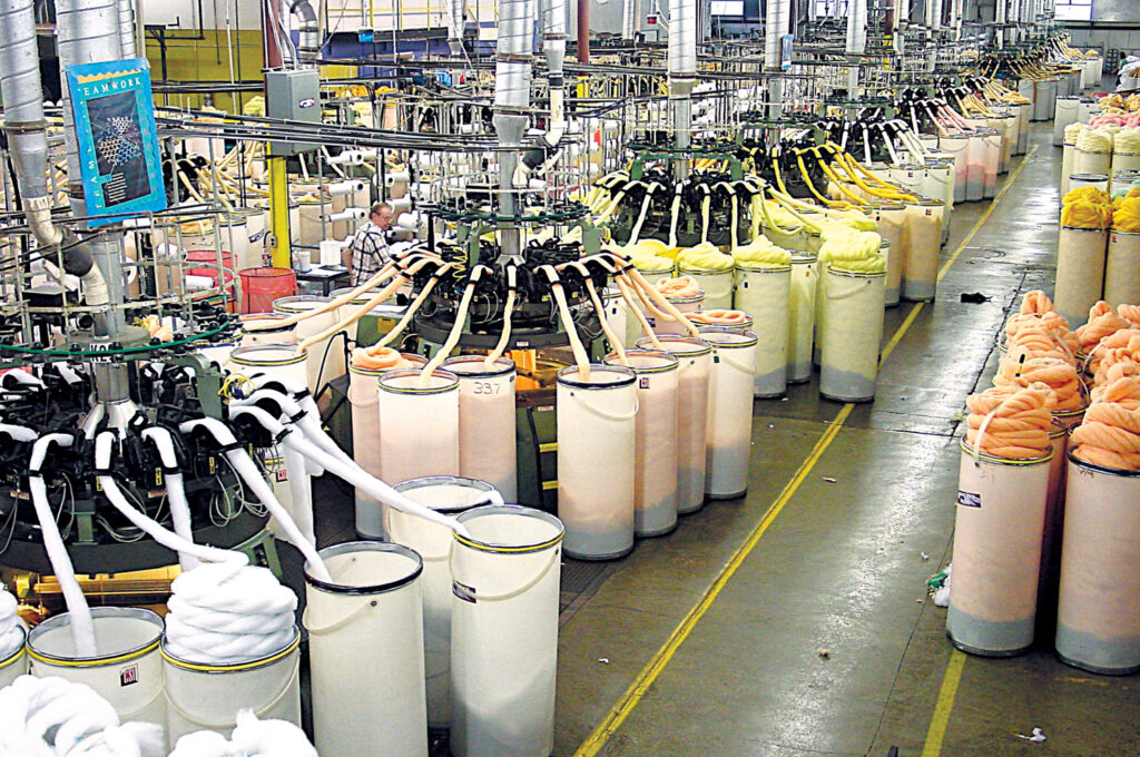 Aerial view of Monterey Mills' textile factory with numerous machines, colorful fabric slivers in large containers, organized in rows.