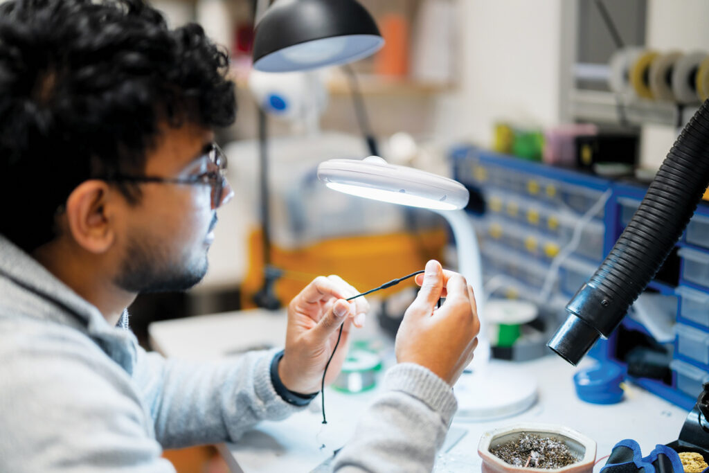 A person with curly hair examines a small object under a bright lamp at a workbench, surrounded by tools and materials.