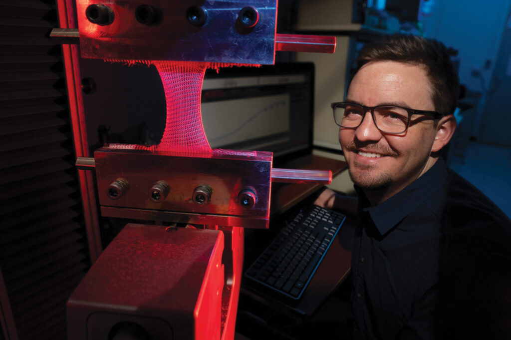 A researcher smiles beside a testing machine, featuring pink synthetic scaffolds under a red light, with a computer in the background.