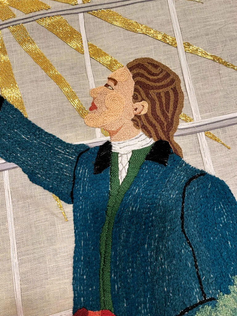 A detailed embroidered depiction of a historical figure in a blue coat with a white collar, reaching upward amid golden rays.