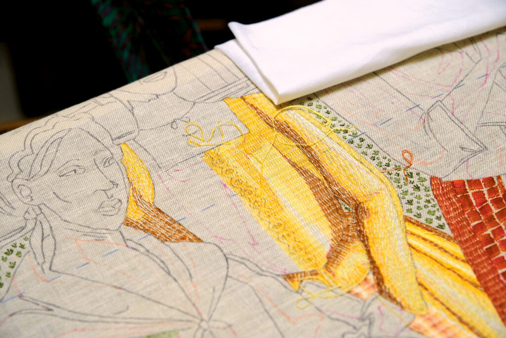Detailed textile artwork featuring colorful stitching of women in traditional attire, with a white fabric corner draped over the edge.