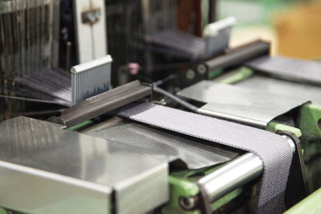 Close-up of a textile loom at OTEX Specialty Narrow Fabrics with gray fabric webbing being woven for a fall-protection harness, which contains Nomex and Kevlar fiber.