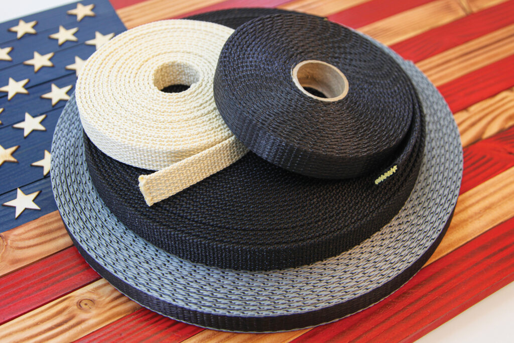 Three rolls of woven webbing from OTEX Specialty Narrow Fabrics in black, navy, and beige are arranged on a textured wooden background with red, white, and blue stripes and stars representing the American flag.