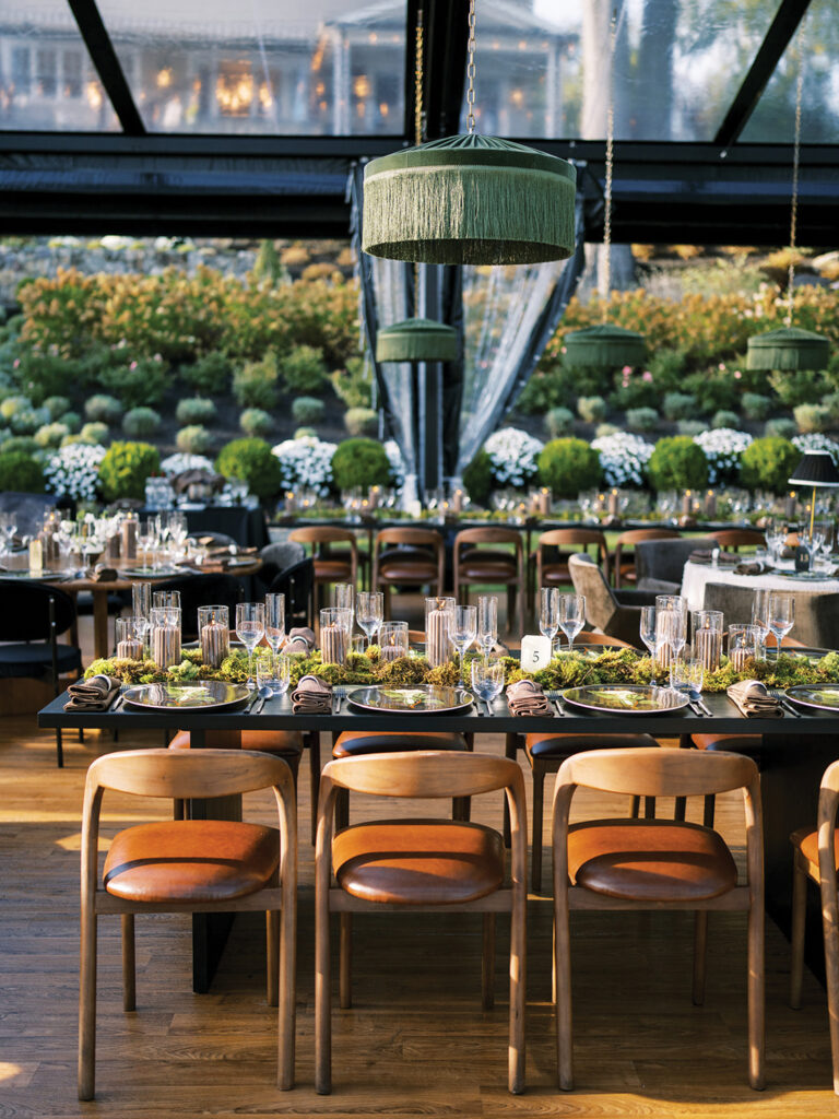 Elegant outdoor dining setup with wooden tables adorned with greenery, glassware, and hanging green lamps, surrounded by lush plants. 