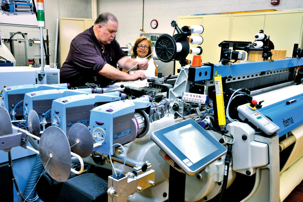 A technician and assistant work at a textile machine, focusing on intricate mechanics. Blue and gray equipment dominates the scene.