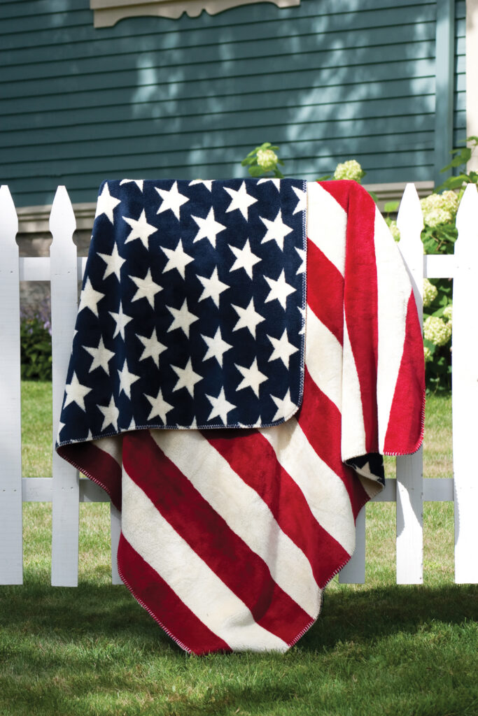 A Denali-brand throw blanket by Monterey Mills, featuring the American flag, with navy blue stars and red and white stripes, draped over a white picket fence.