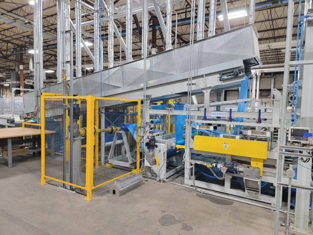 01-Menzel-36-In-Kiss-Coating-Line Yellow safety cage surrounds a blue and gray coating line with layered conveyor systems, set within a spacious industrial environment.